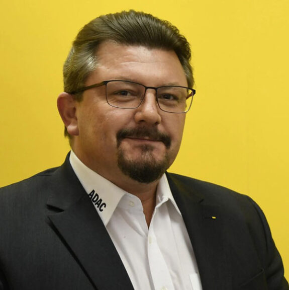 Jürgen Hildebrandt vor einer gelben Wand. Er trägt Brille, einen Anzug mit weißem Hemd und Bart.